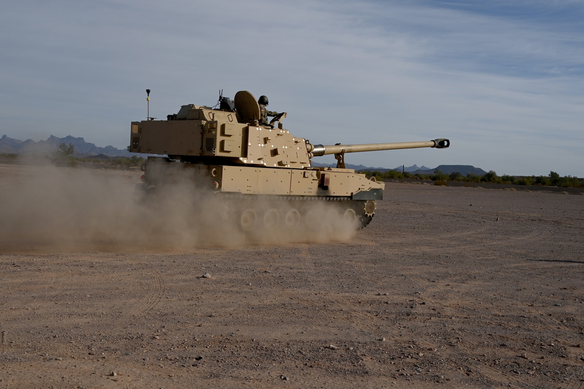 A crew at Yuma Proving Ground works around a large cannon barrel test article on an outdoor test pad, with the mountainous desert terrain in the background