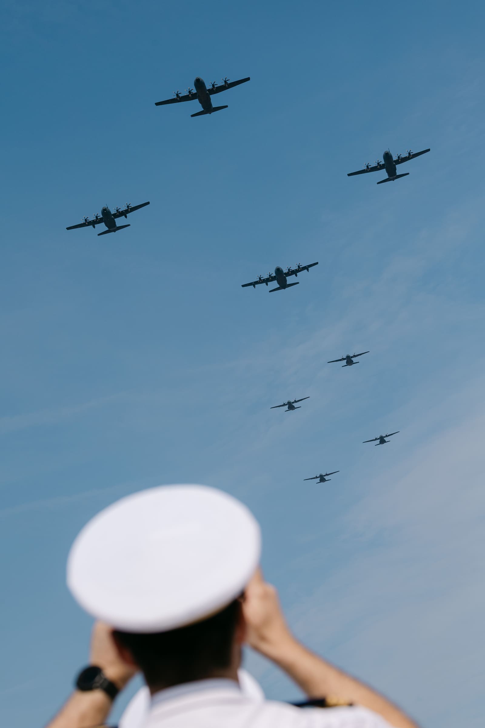 A formation of eight French Air and Space Force transport aircraft flies overhead in a V formation during the 2026 Bastille Day flypast, viewed from below with a saluting naval officer in a white cap in the foreground