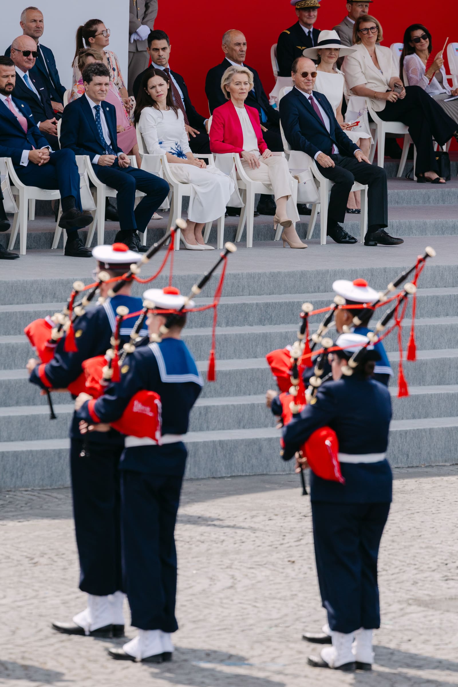 Republican Guard bagpipers in navy-blue uniforms with red-tasseled bagpipes march past the presidential tribune at the 2026 Bastille Day parade, with seated dignitaries including European Commission President Ursula von der Leyen visible behind them