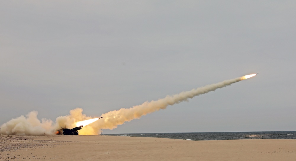 A HIMARS wheeled rocket launcher firing during a NATO training exercise in Poland, with a rocket visible leaving the launch tube